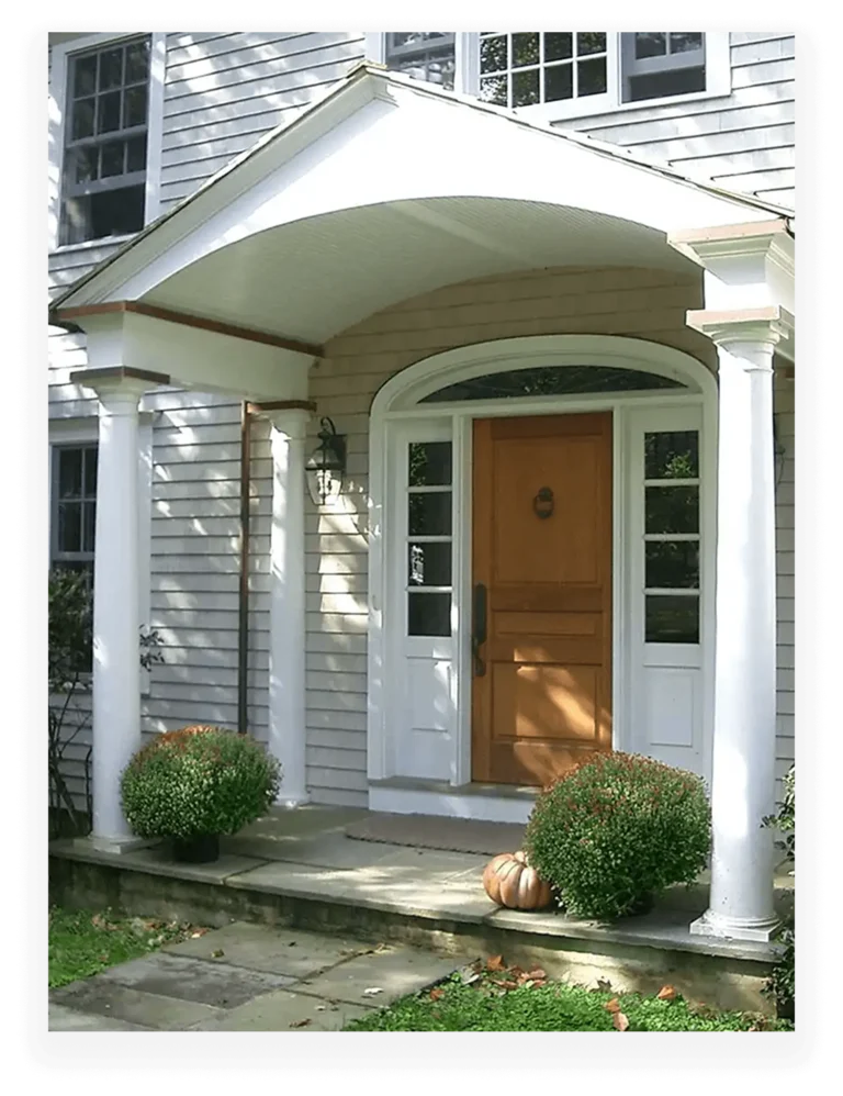 A home’s front entrance with a wooden door flanked by sidelights, a curved portico supported by white columns, and decorative shrubs on the porch.